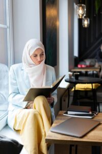 A young Muslim woman in a hijab reading a book at a modern cafe, with a laptop on the table.